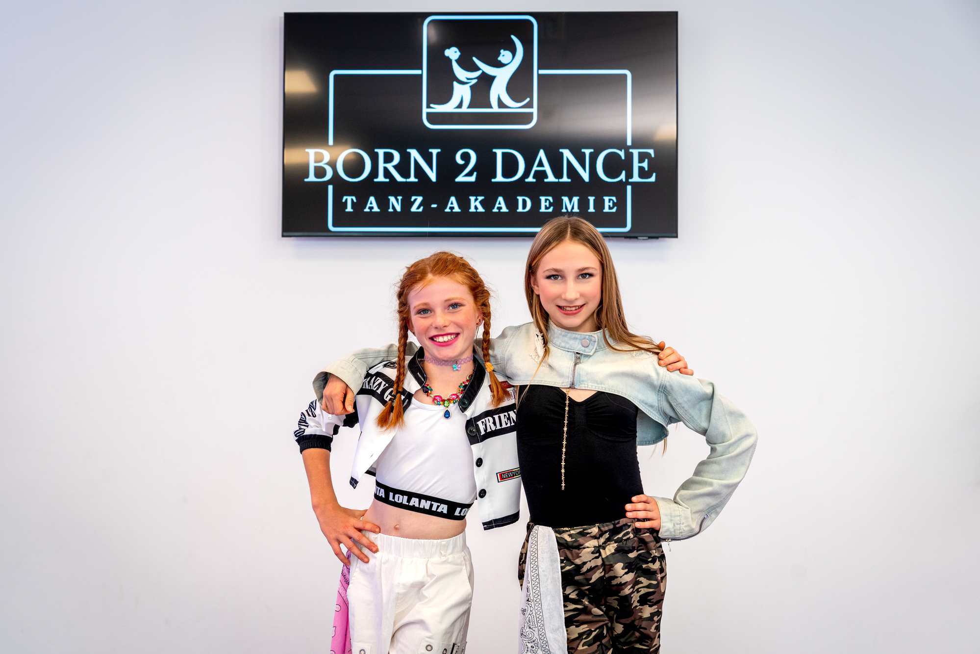 Zwei junge Tänzerinnen posieren lächelnd vor dem Born 2 Dance Tanz-Akademie Schild im Studio